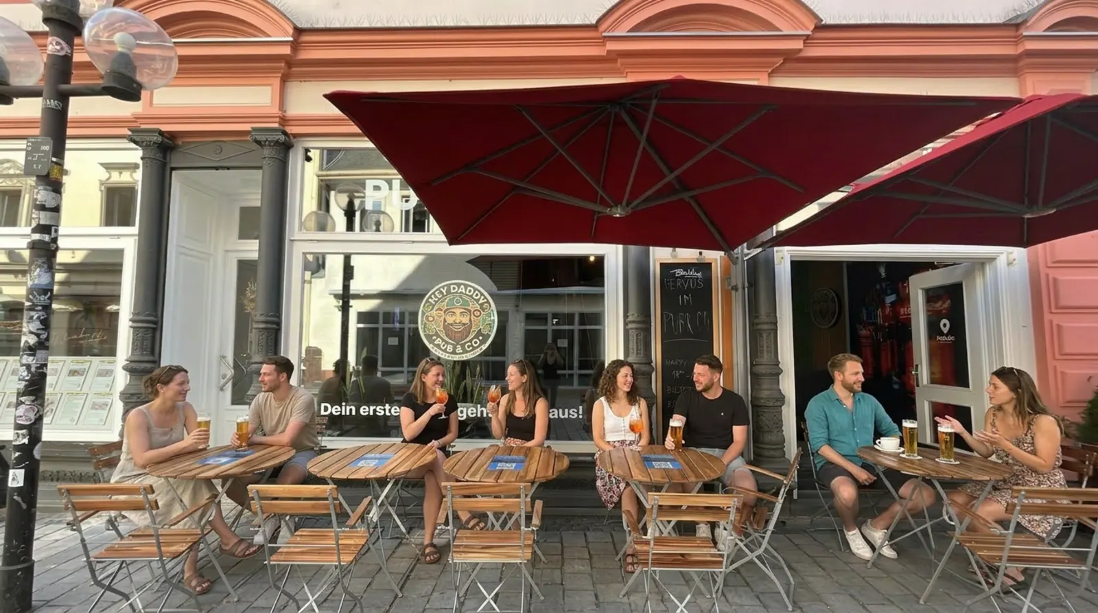 Pub&Go Bayreuth Außenterrasse mit Gästen unter roten Sonnenschirmen